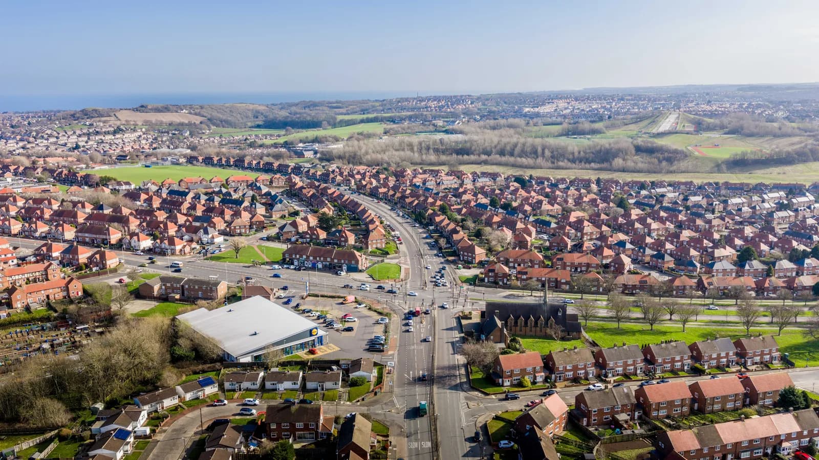 An aerial view of a residential area with a large road passing through, surrounded by houses with red roofs and green spaces, under a clear sky.