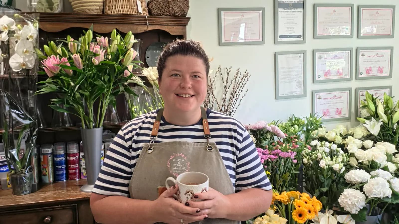 A smiling florist stands in her shop holding a mug, surrounded by various fresh flowers and certificates on the wall.