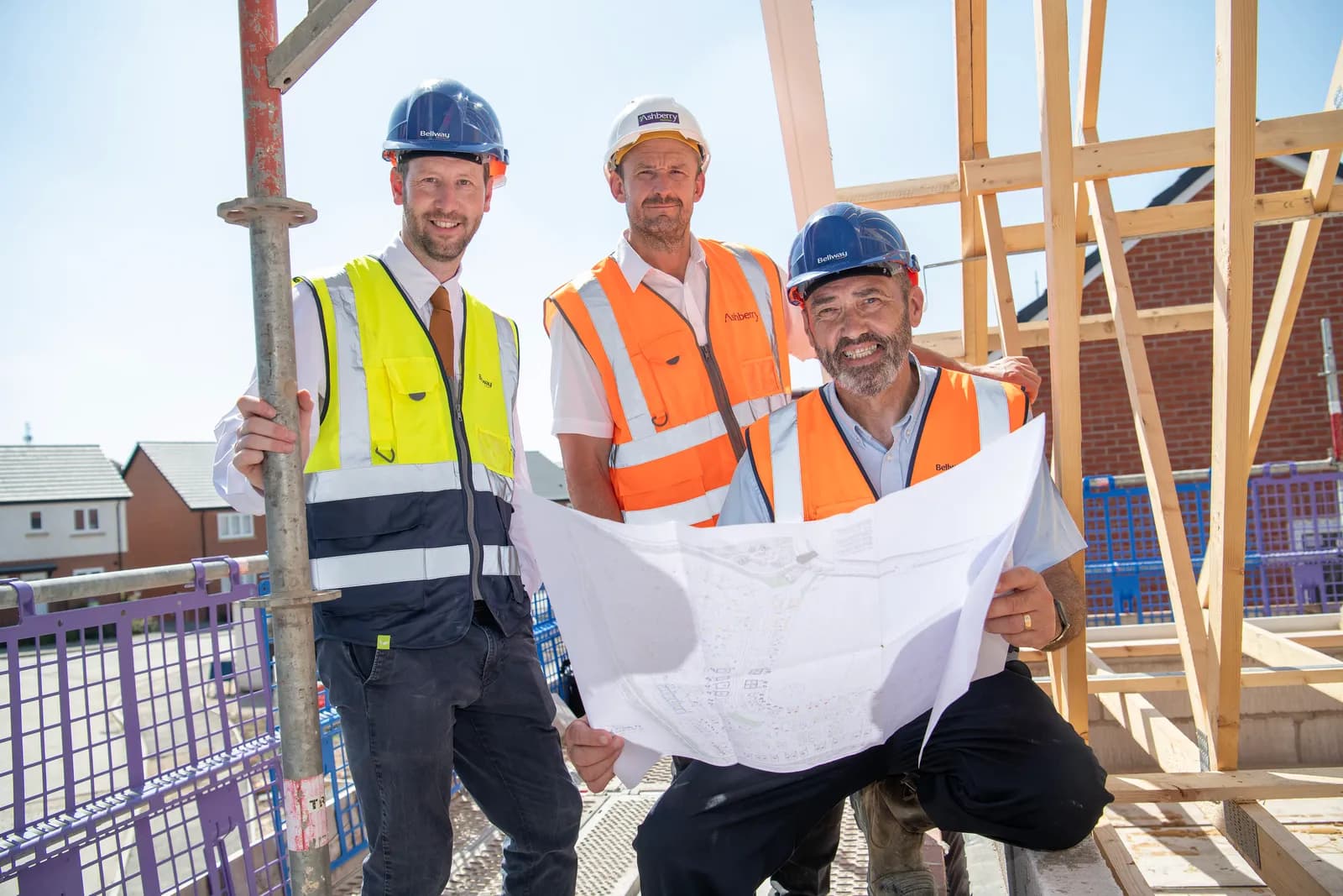 Three construction professionals wearing hard hats and high-visibility vests smile at the camera while holding architectural plans on a construction site.
