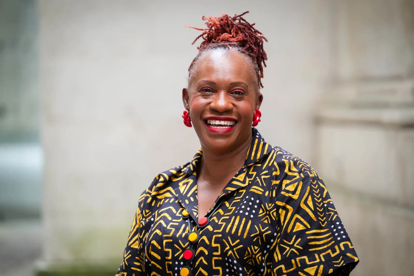 A smiling woman with red locs and bright red lipstick wears a patterned yellow and black shirt and red earrings.