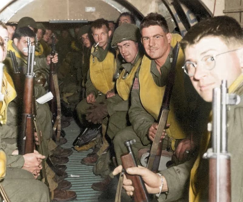 A group of soldiers in military uniforms and yellow life vests sit in a dark aircraft cabin, holding rifles.