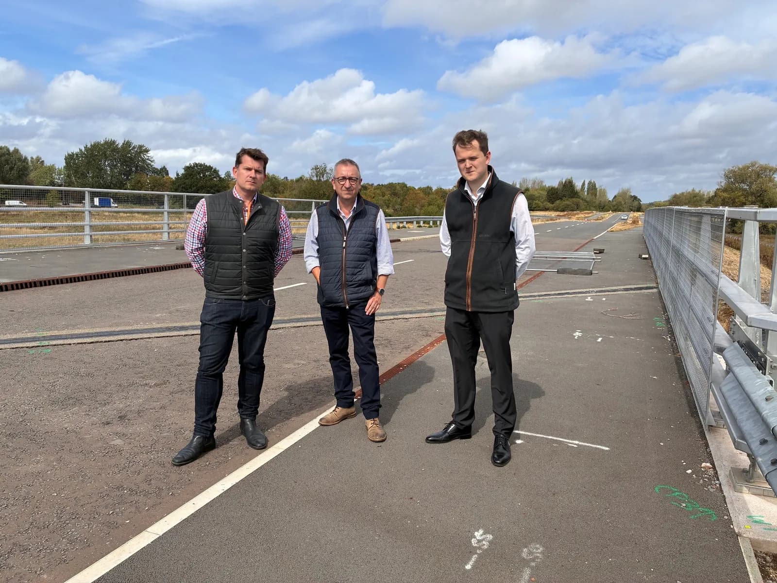 Three men stand on a partially constructed road, with one wearing a plaid shirt and vest, one wearing a light blue shirt and vest, and one wearing a white shirt and black vest.