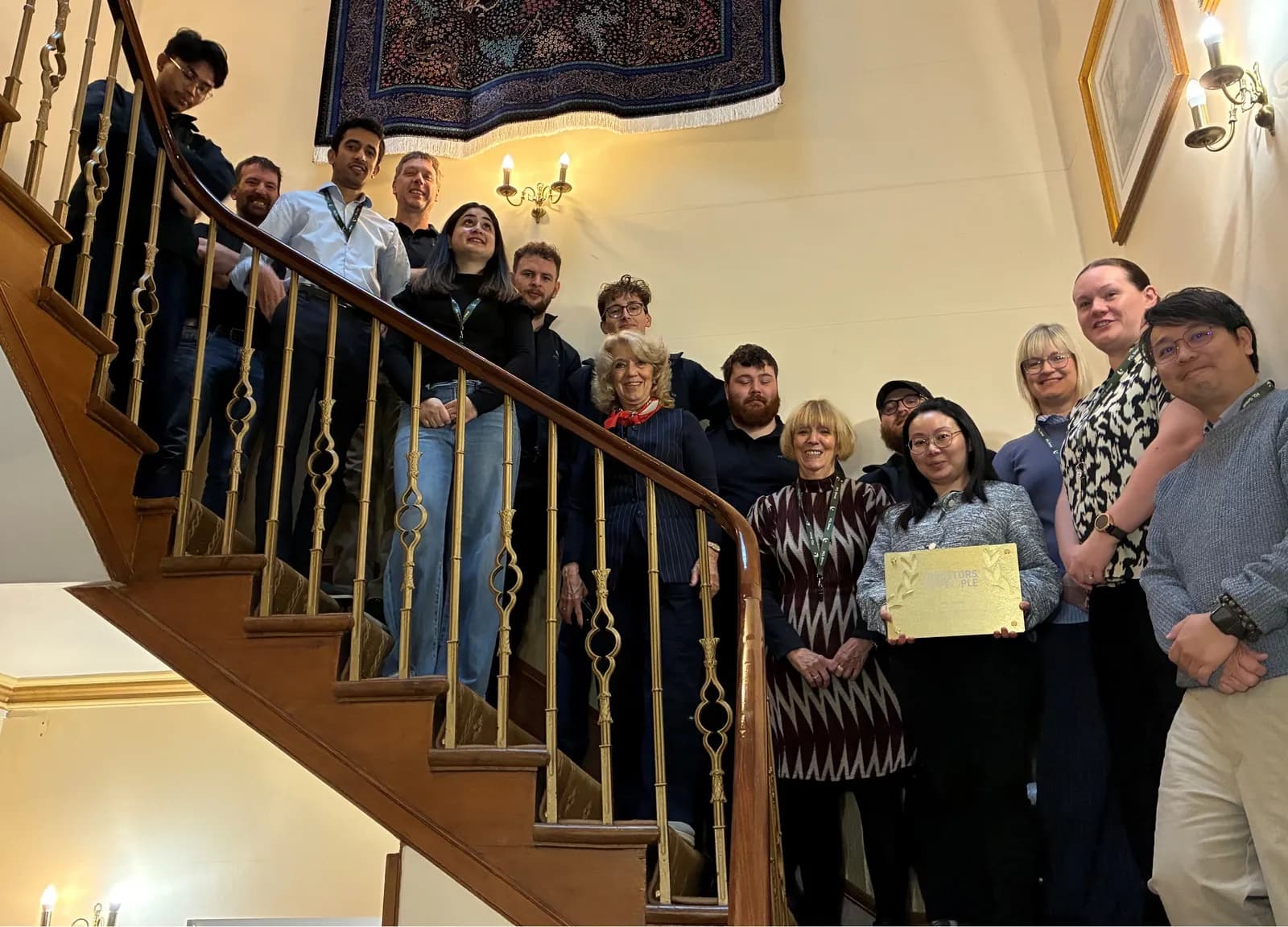 A diverse group of people, some smiling, are gathered on a grand wooden staircase with a golden railing.