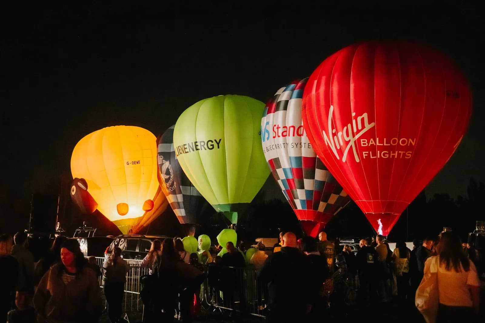A row of brightly illuminated hot air balloons, including one red with 'Virgin Balloon Flights' written on it, another green with 'JEBENENERGY', and a yellow one, stand at night with a crowd of people gathered around them.