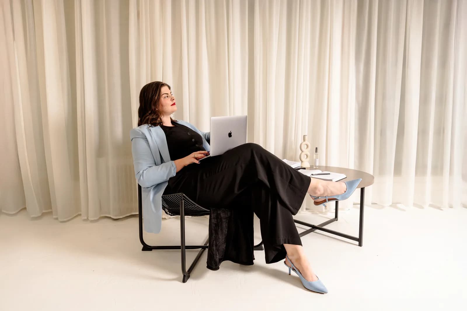 A woman in a light blue blazer and black clothing sits with a laptop on her lap, looking upwards and to the right, against a background of white curtains.