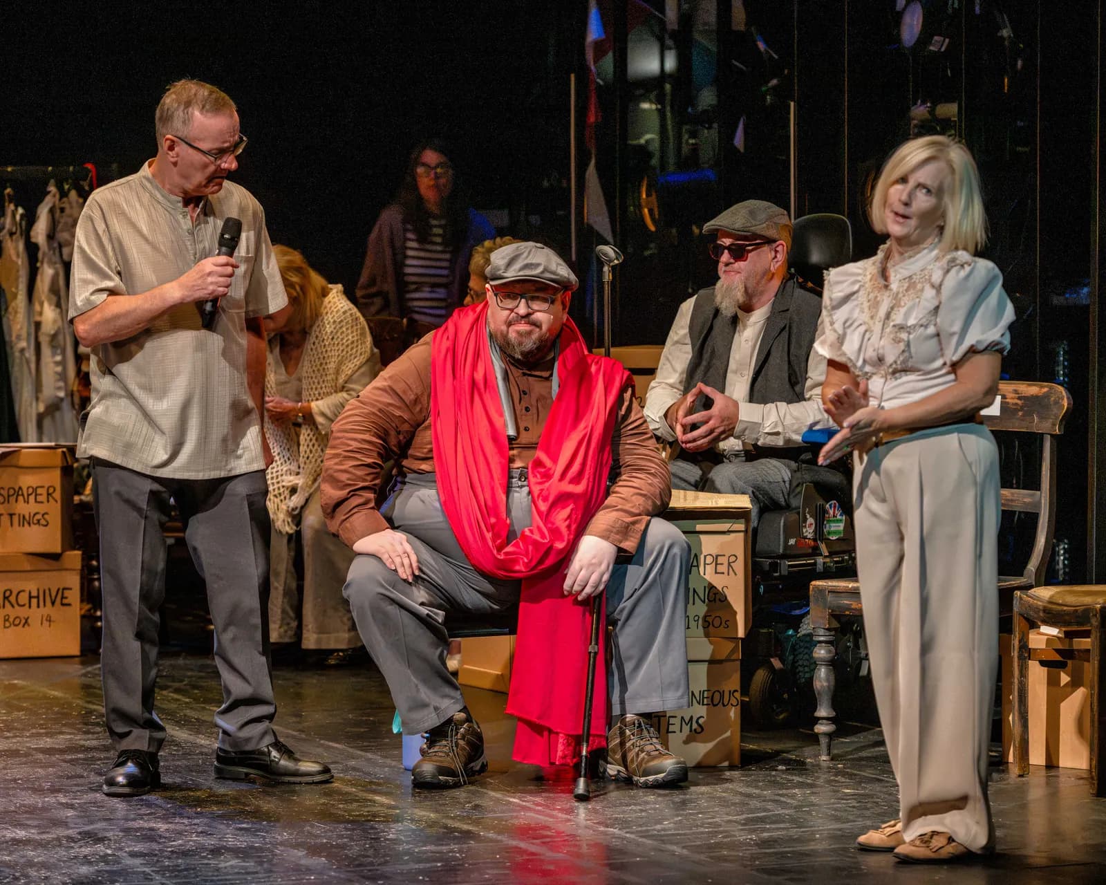 A man in glasses holding a microphone talks with three people seated and a woman standing in a theatrical setting with props.