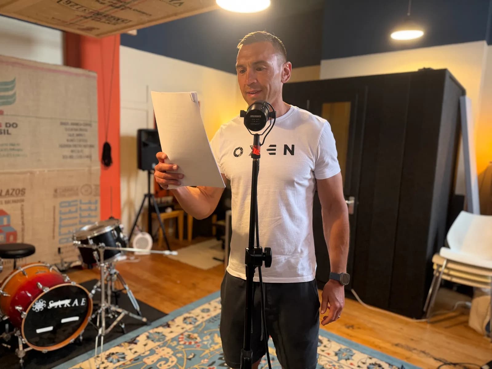 A man in a white t-shirt and black shorts reads from a piece of paper into a microphone in a room with a drum set and a sound booth.