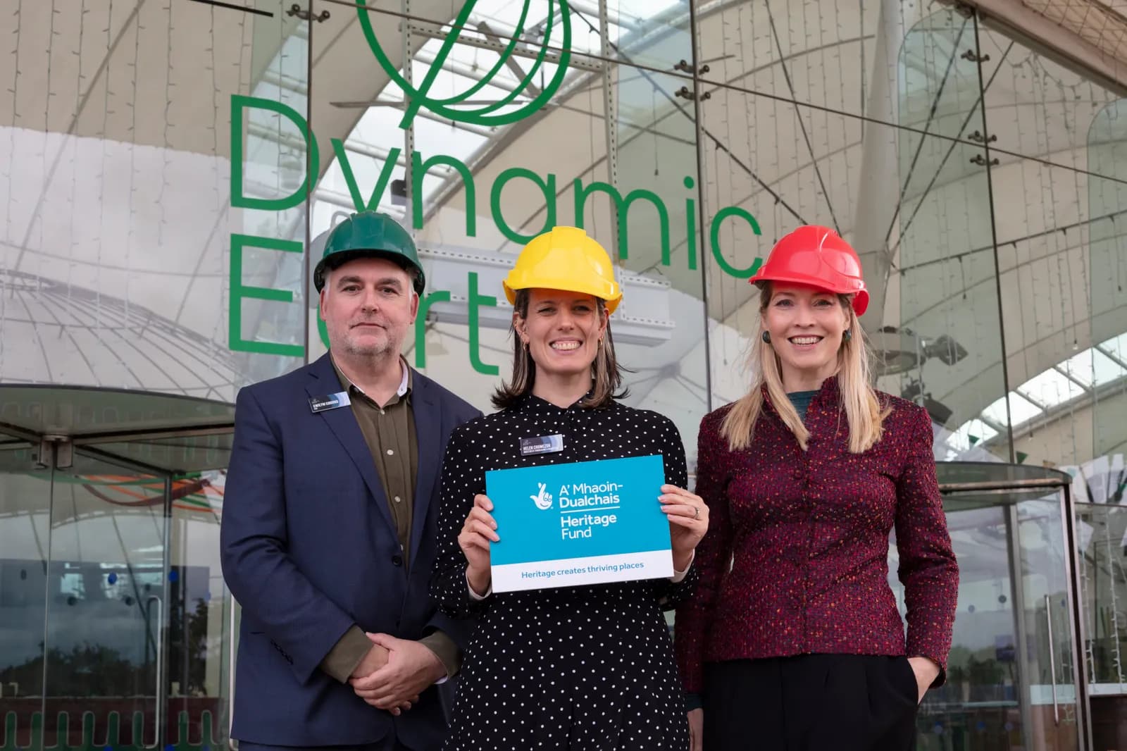 Three people wearing hard hats, one man and two women, stand in front of a glass building with green text that reads "Dynamic Earth," and the woman in the center holds a blue sign that says "A' Mhaoin-Dualchais Heritage Fund."