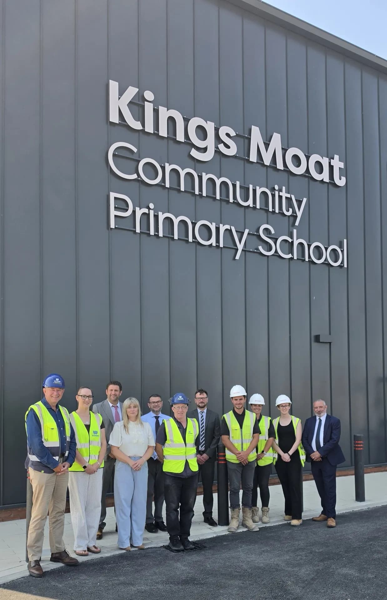 A group of ten adults, some wearing construction vests and hard hats, stand smiling in front of the Kings Moat Community Primary School building.