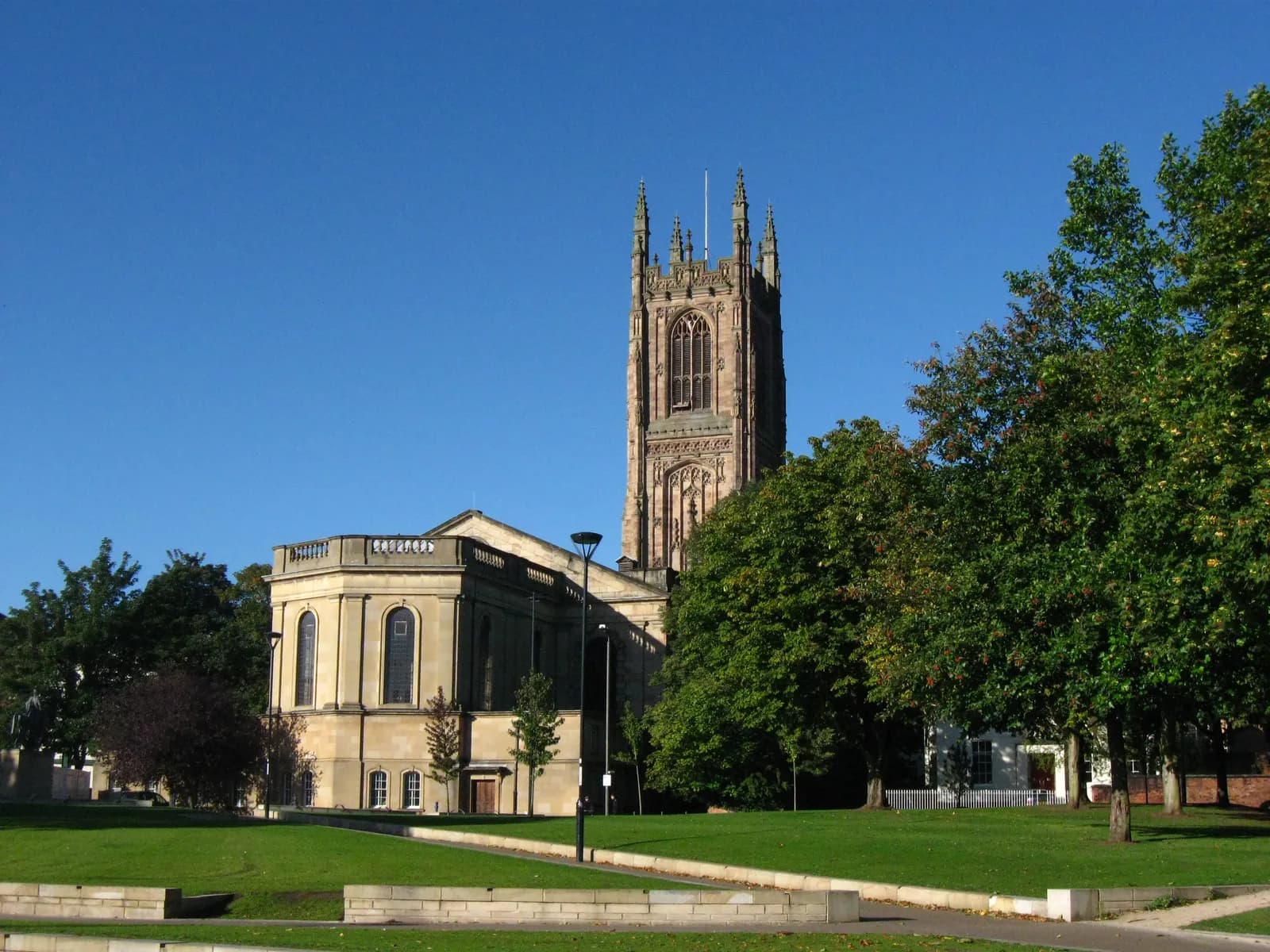 A historic stone church with a tall, ornate tower stands prominently on a sunny day, surrounded by green grass and lush trees under a clear blue sky.