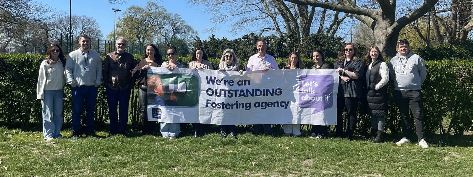 A group of people, standing outside on a sunny day, holding a banner that reads "We're an OUTSTANDING Fostering agency. Let's talk about it."