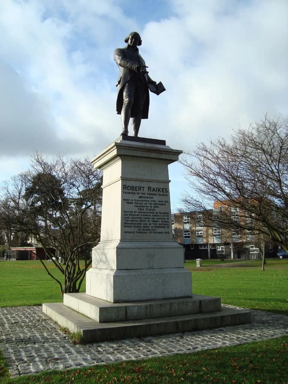 A towering stone statue of Robert Raikes in a park with green grass and trees, with a building in the background.