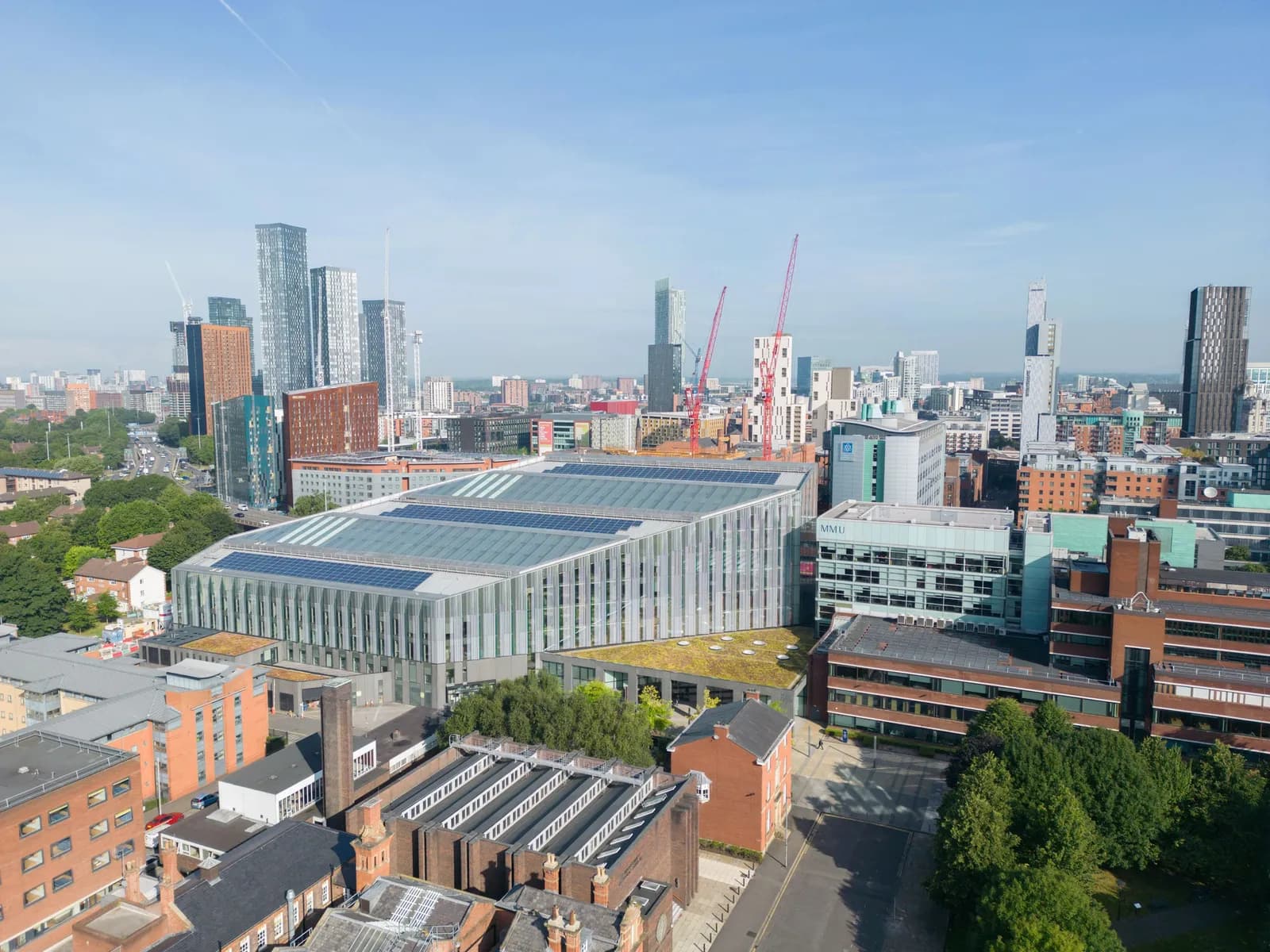 An aerial view showcases a modern, glass-paneled building with solar panels on its roof, surrounded by other city buildings and distant skyscrapers under a clear blue sky.