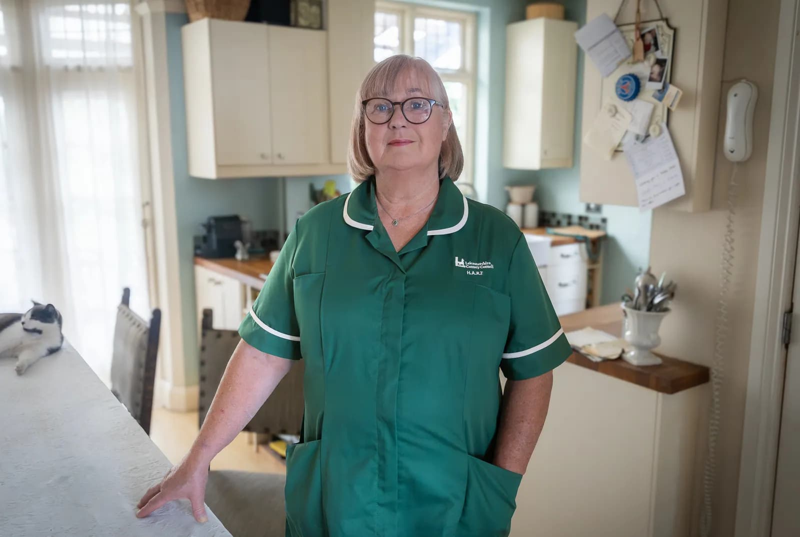 A woman in a green medical uniform with white trim stands in a kitchen, looking directly at the camera.