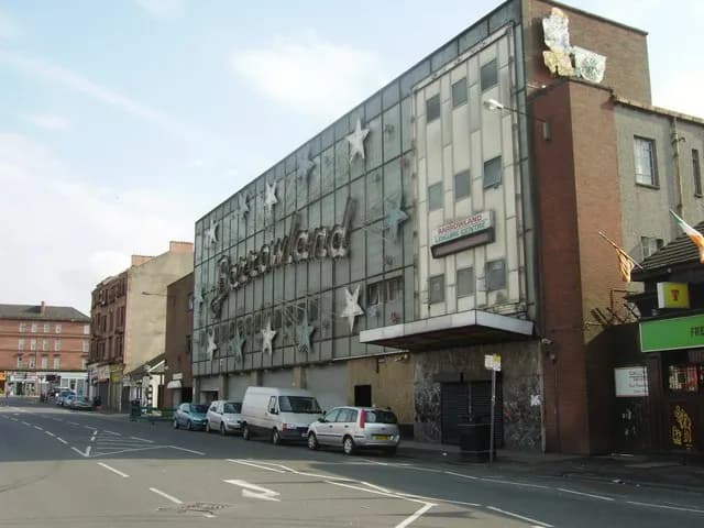 The Barrowland Ballroom, a large building with windows and stars on its facade, stands prominently on a street lined with other buildings and parked cars.