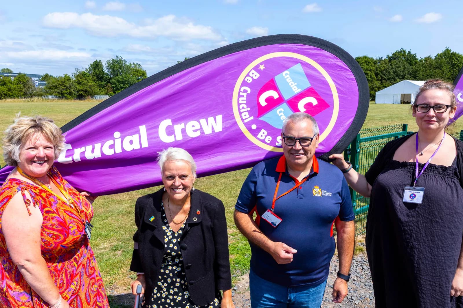 Four adults, two women and two men, standing outdoors and holding a large purple banner that reads "Crucial Crew" and features a circular logo.