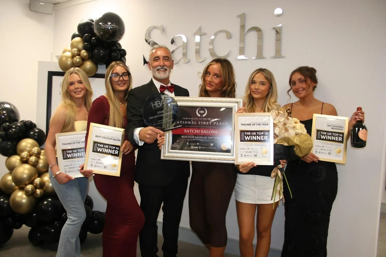 A man in a suit and bow tie stands with five women, all holding awards and certificates in front of a wall with metallic lettering spelling "Satchi."