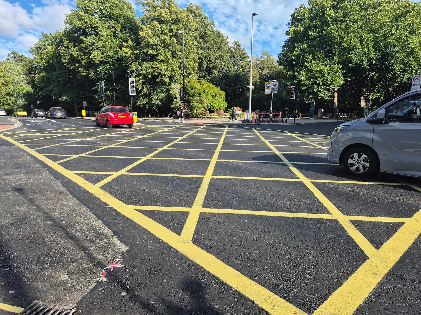A newly painted yellow grid pattern covers an asphalt intersection with cars stopped at traffic lights and green trees in the background.