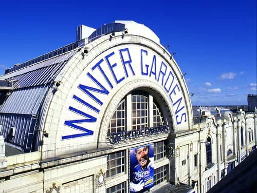 The white facade of the Winter Gardens building features blue block letters spelling out its name in an arch over a large window, with a poster of a clown visible below.