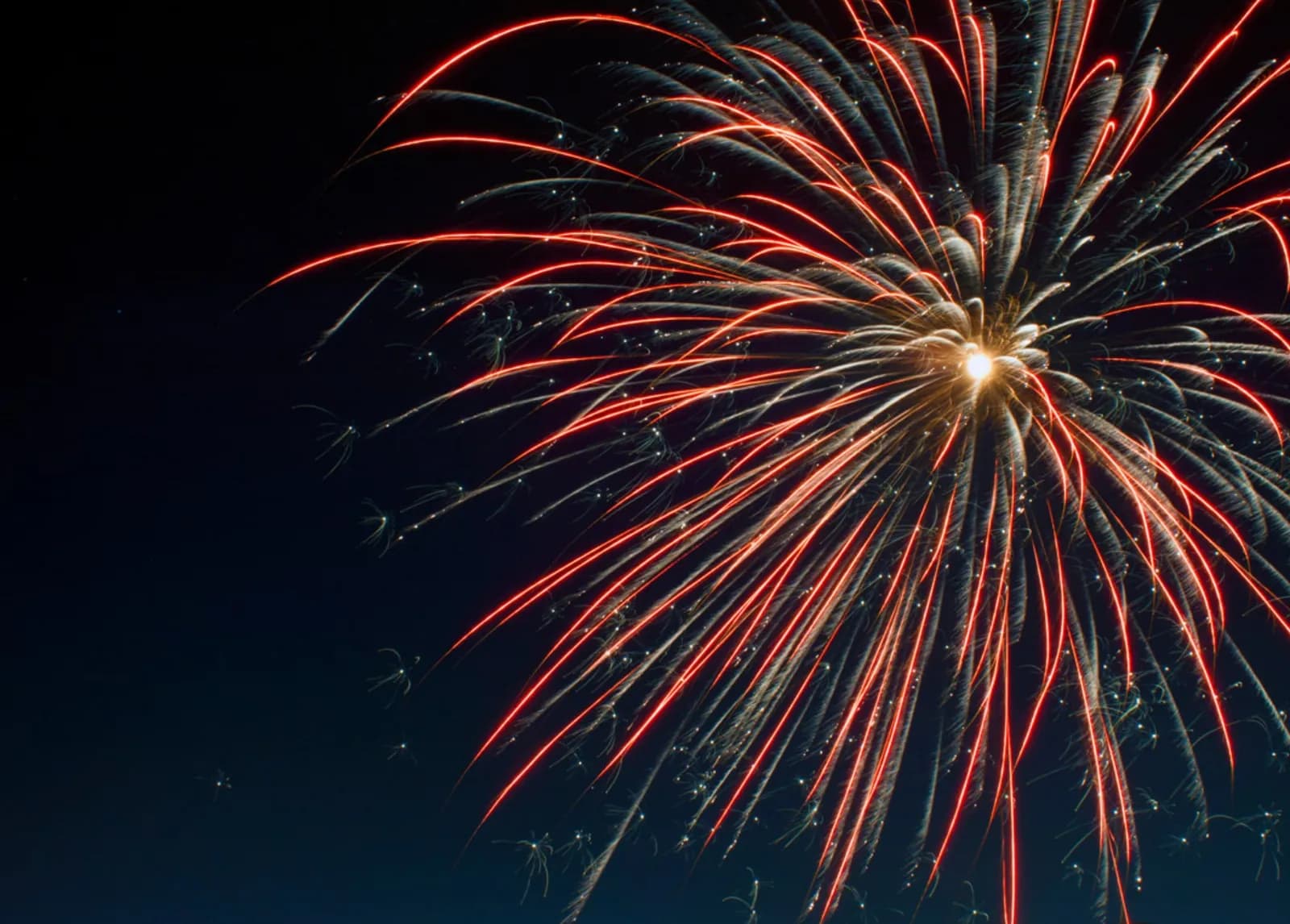 Red and white fireworks burst against a dark night sky, with trails of light extending outwards from a bright central explosion.