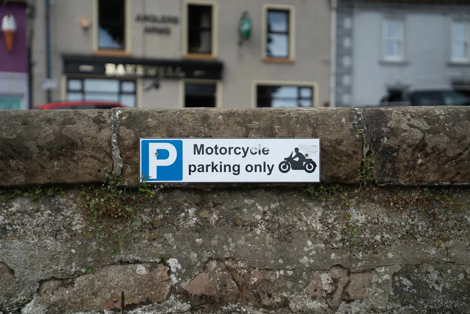 A white sign with a blue P for parking and text "Motorcycle parking only" with a motorcycle icon is affixed to a stone wall, with blurred buildings in the background.