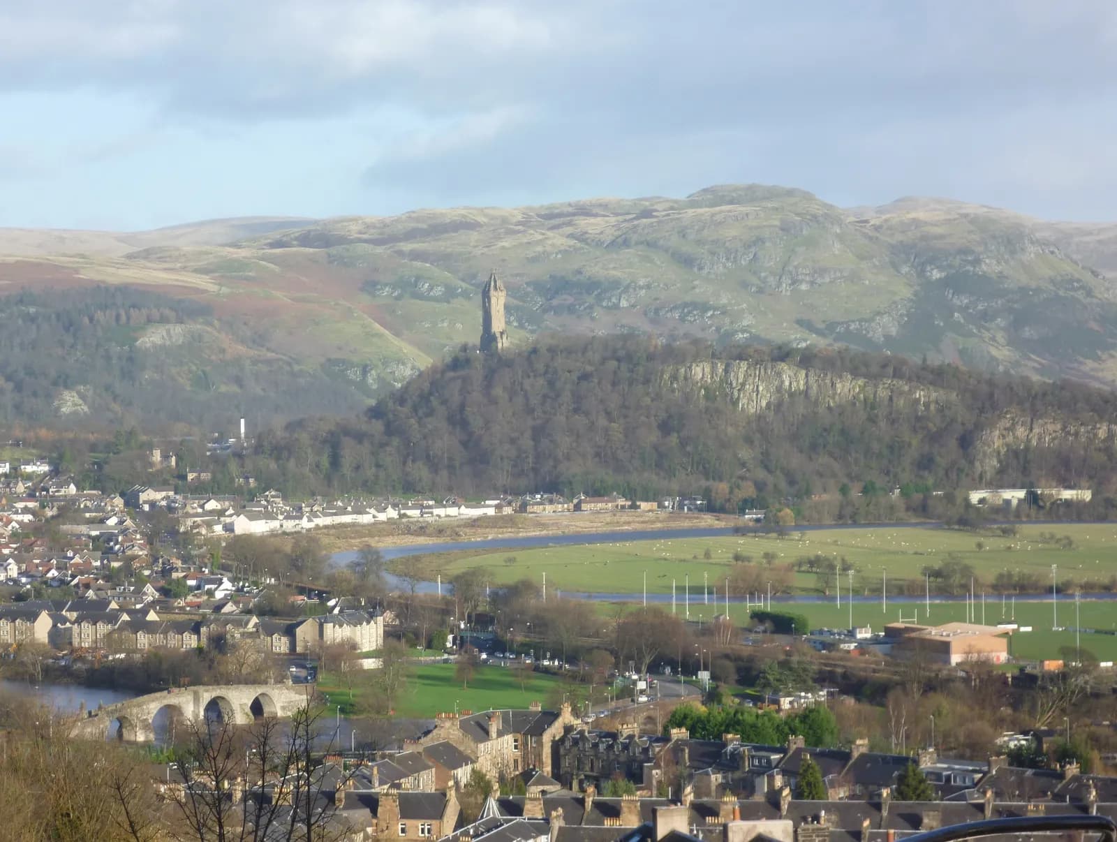 A scenic view of a town with a river flowing through it, a stone arch bridge, and a prominent tower on a wooded hill in the background, set against rolling hills under a cloudy sky.
