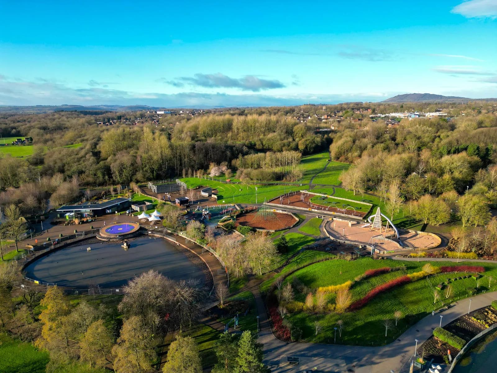 An aerial view of a large park with a lake, playgrounds, buildings, and vast green spaces surrounded by trees under a bright blue sky.