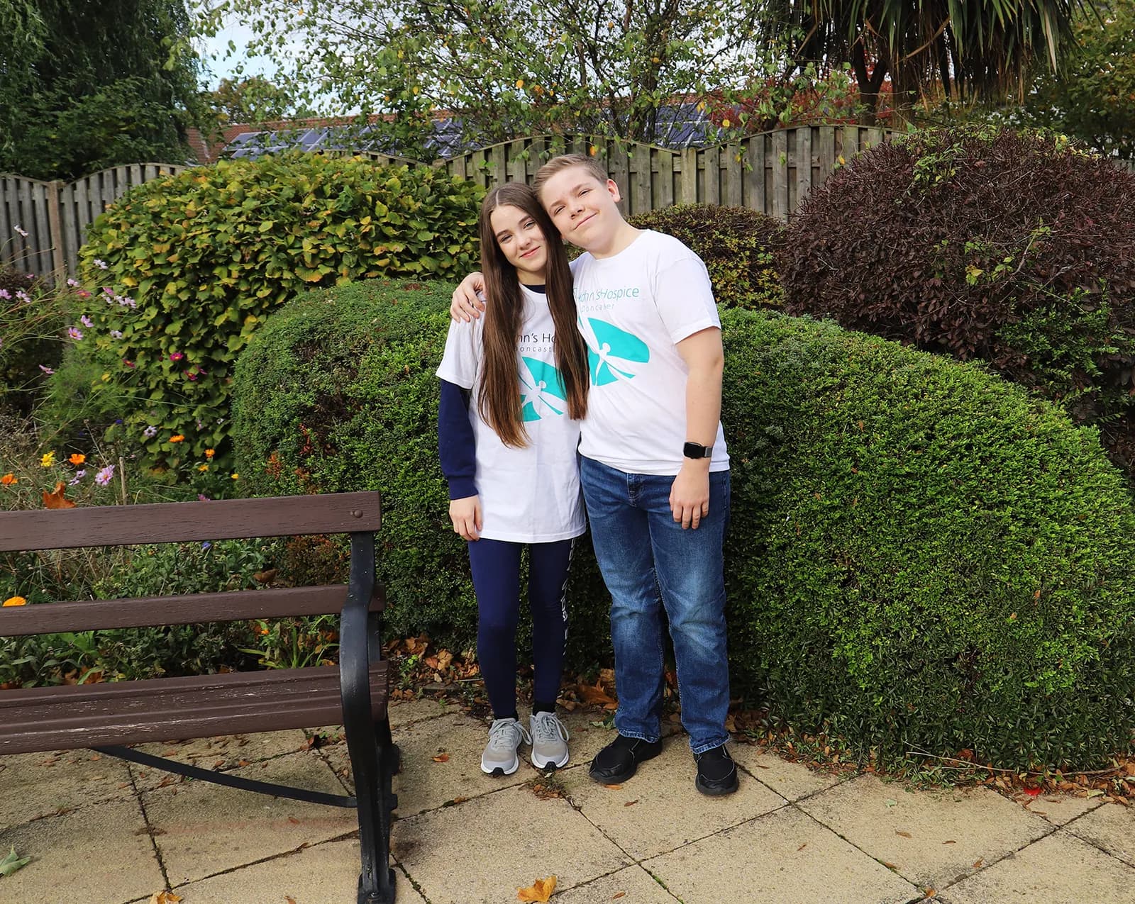 A girl with long dark hair and a boy with short blonde hair stand side-by-side, smiling with their arms around each other, wearing matching white t-shirts with a light blue logo, in a garden with green bushes and a wooden fence.