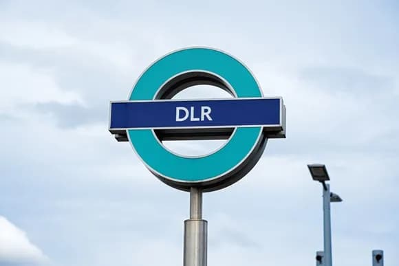 A close-up shot of a London DLR sign, a circular teal and white emblem with a blue bar across the middle that reads "DLR" in white capital letters, set against a cloudy sky.