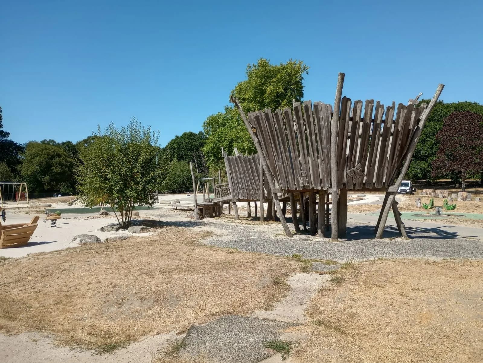 A large, wooden play structure resembling a boat or fort stands in a sandy playground under a clear blue sky, surrounded by green trees and dry grass.