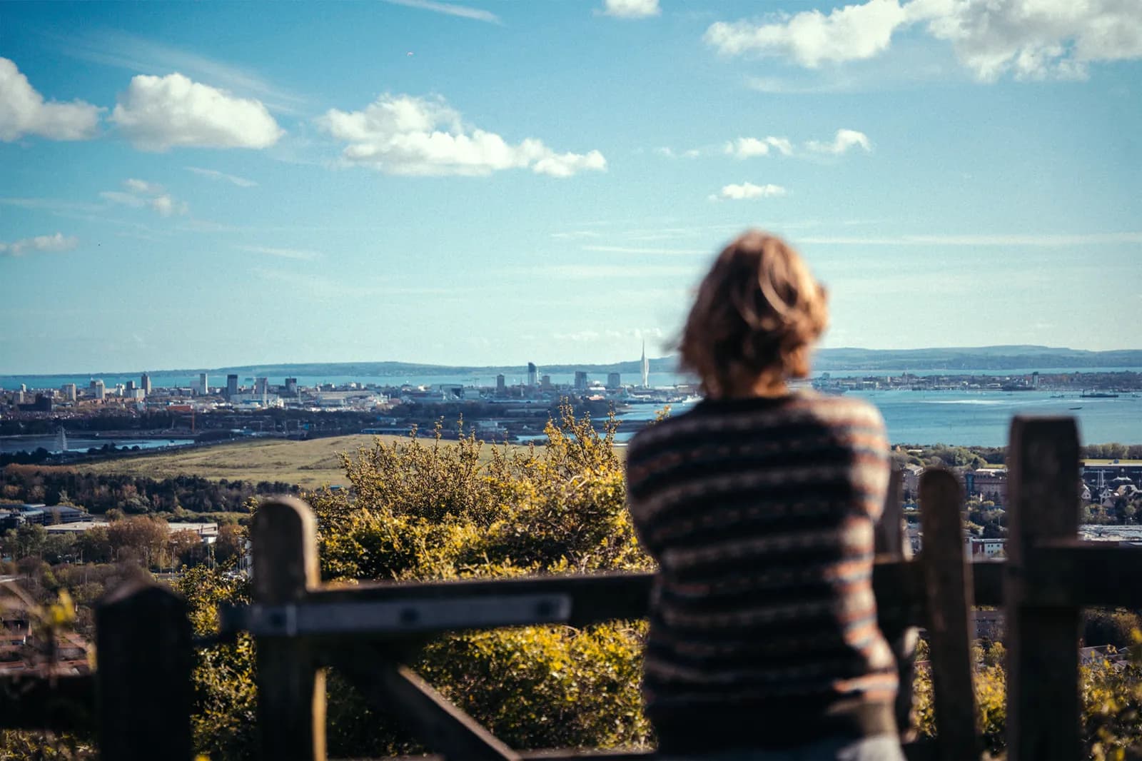 A person with short, curly brown hair and a striped sweater stands with their back to the viewer, looking out over a city skyline next to a body of water under a blue sky with scattered clouds.