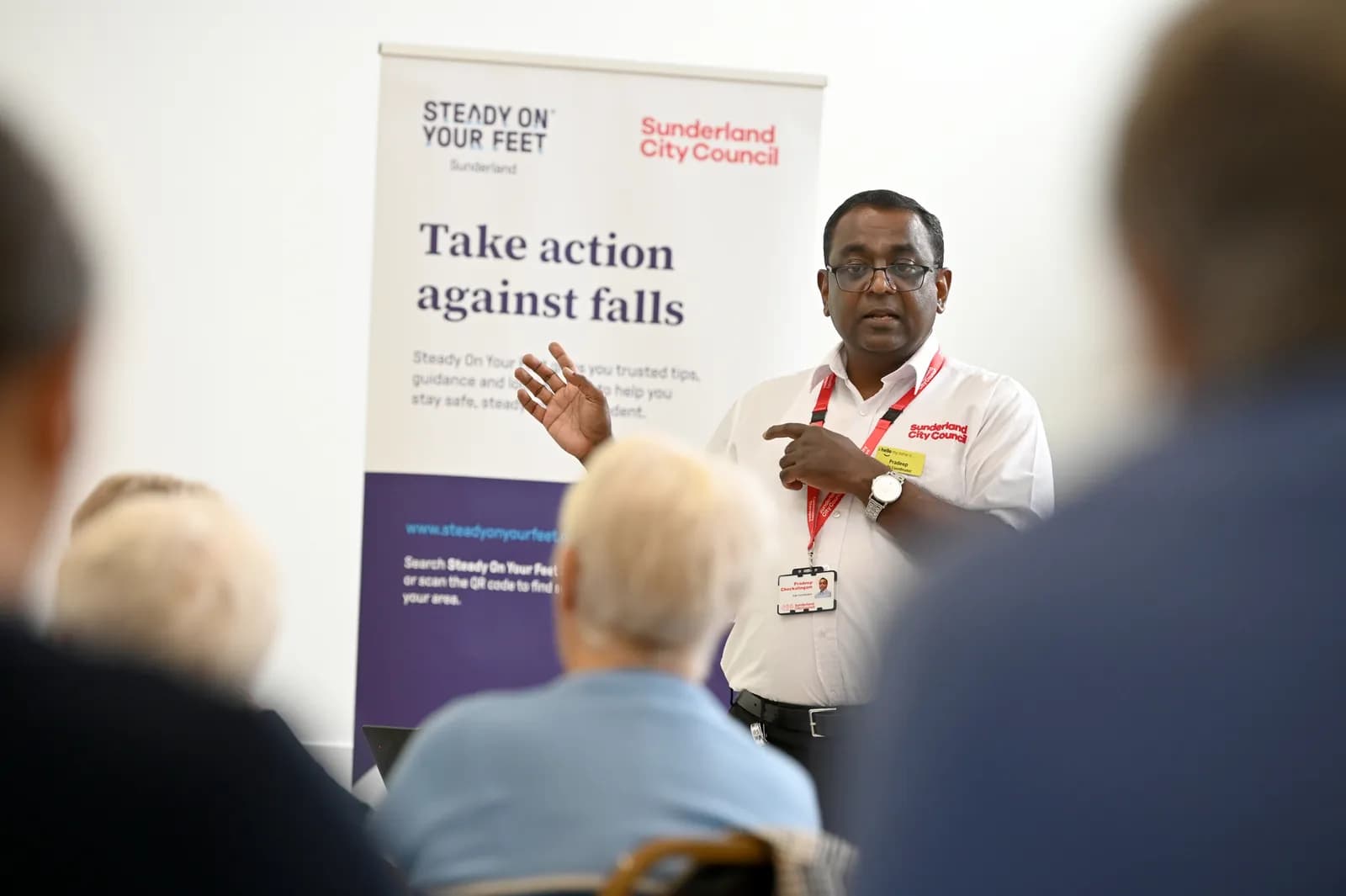 A man in glasses wearing a white shirt with an ID badge and red lanyard gestures with both hands as he speaks to an unseen audience, standing in front of a banner that reads "Steady On Your Feet, Sunderland City Council, Take action against falls."
