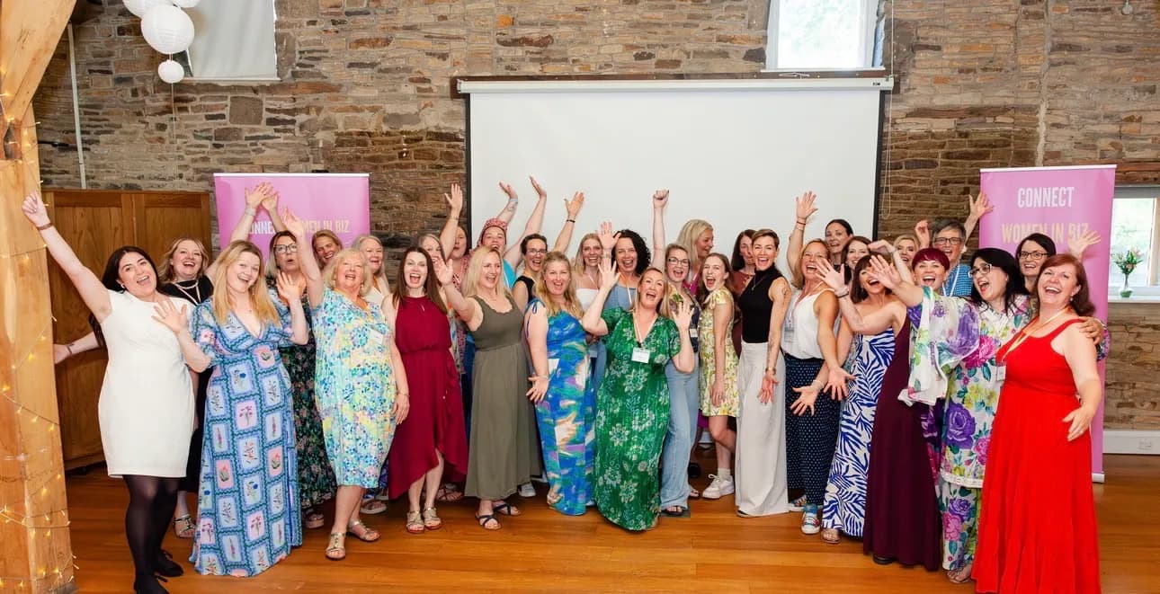 A large group of women celebrating, cheering, and waving their hands in the air at an indoor event, standing in front of a white screen and a stone wall.