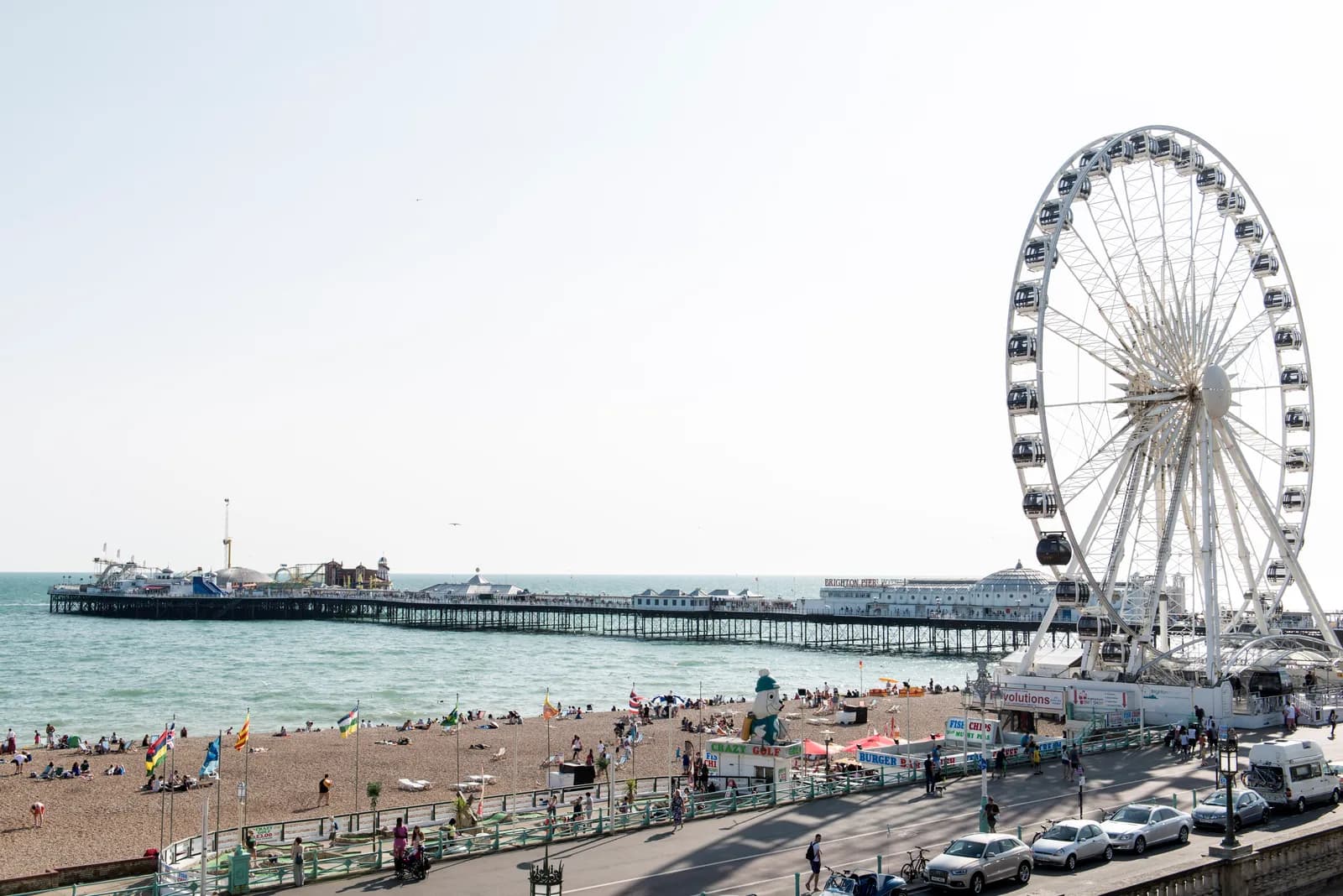 A wide shot of Brighton beach with the iconic pier stretching into the calm sea on the left and a large Ferris wheel dominating the right side of the frame.