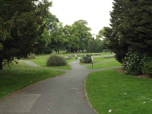 A park scene with paved pathways winding through green grass and surrounded by lush trees, with a few people walking in the distance.