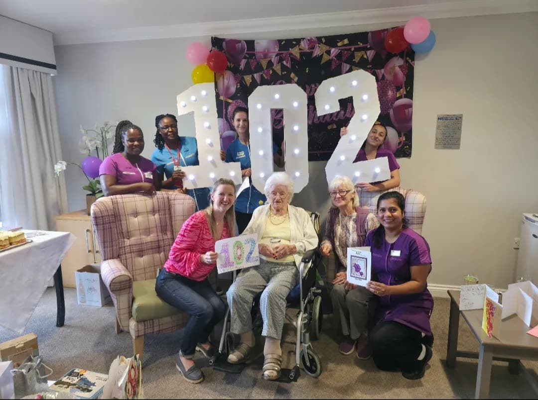 A group of women, including caregivers and elderly residents, smiling and celebrating a 102nd birthday with light-up numbers and balloons.