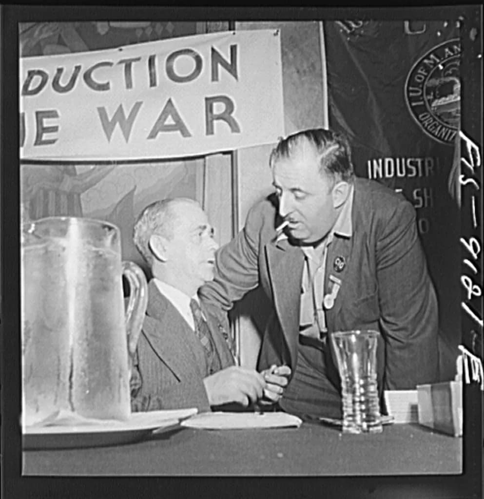 Two men in suits at a table, one leaning in with a cigarette in his mouth, talking to the other. A banner in the background reads "PRODUCTION THE WAR."