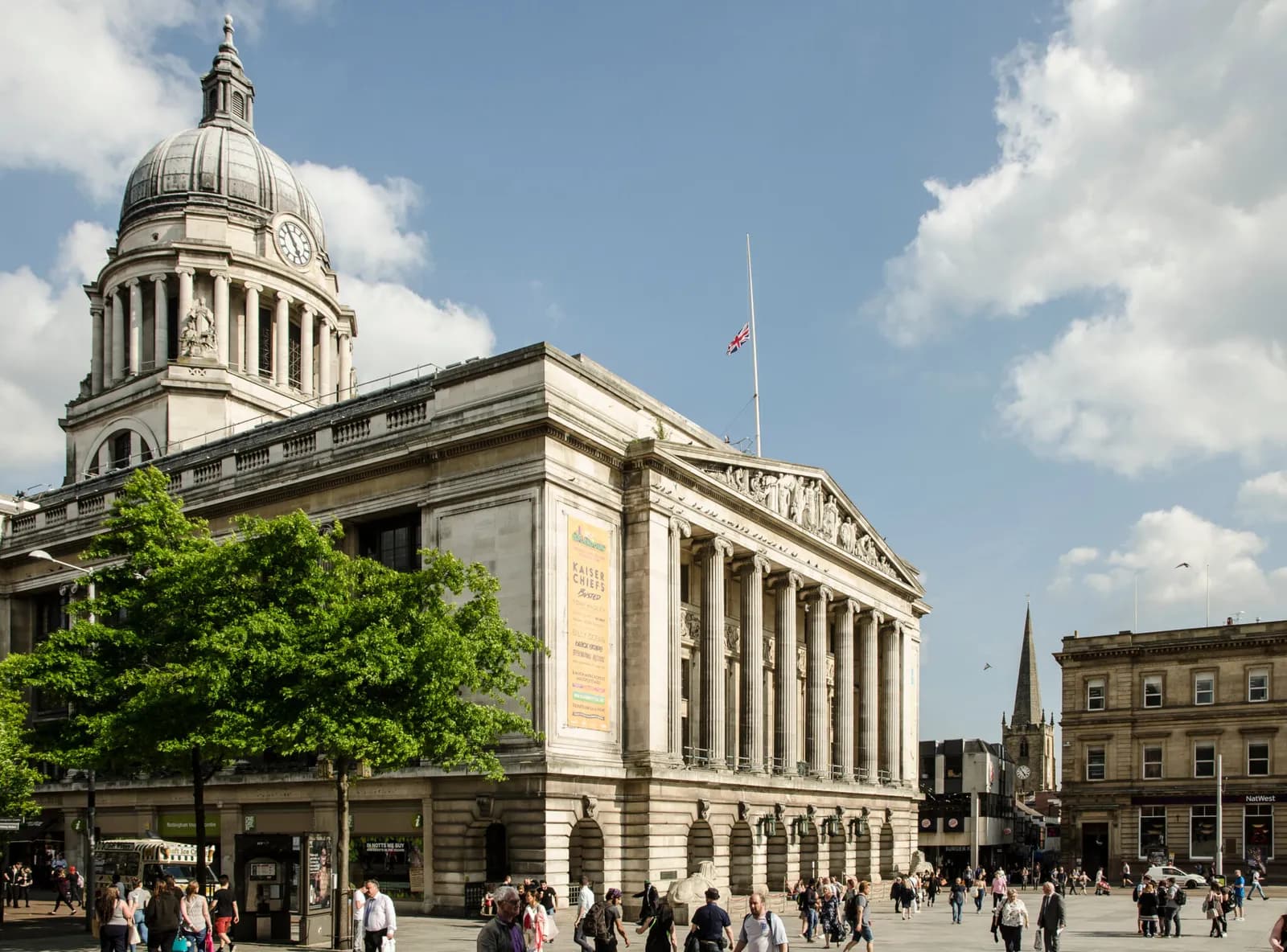 A wide shot of the Nottingham Council House, a grand building with a large dome and clock tower, surrounded by a bustling public square with people walking around.