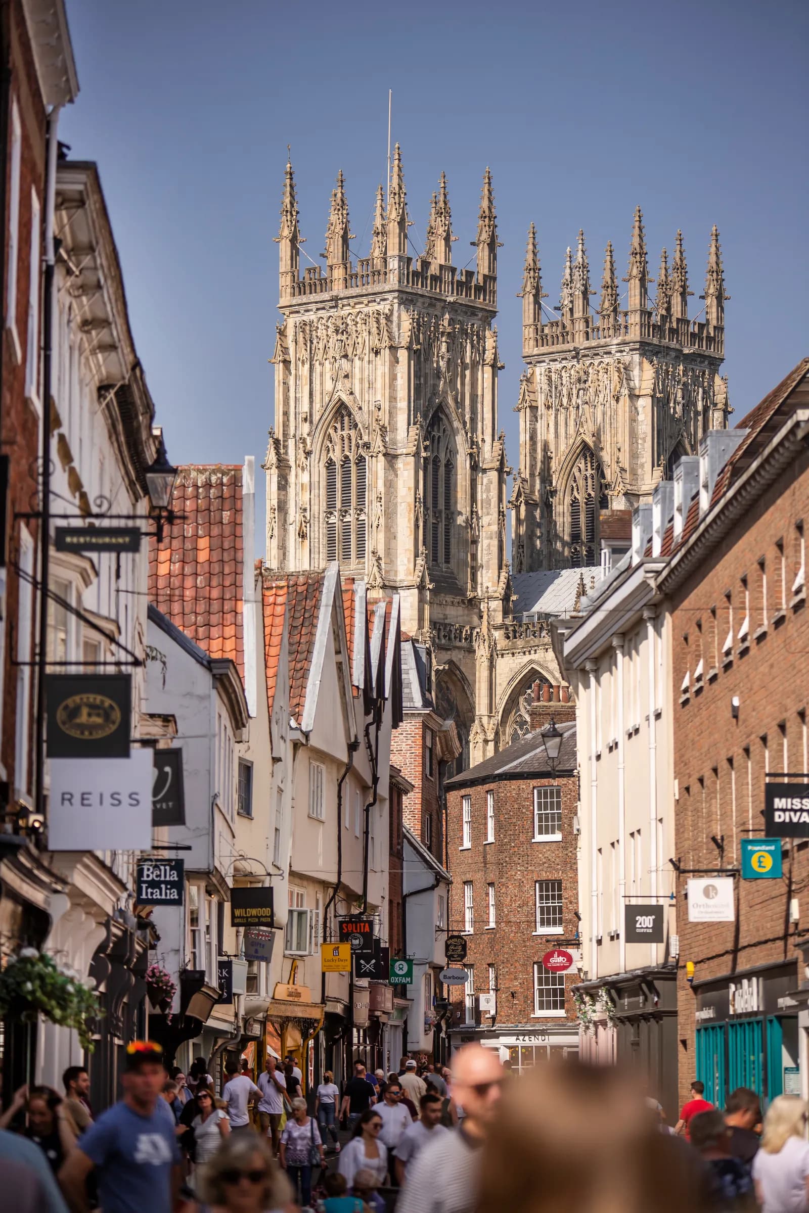 A bustling street lined with shops and people, with the grand towers of a gothic cathedral visible in the background.