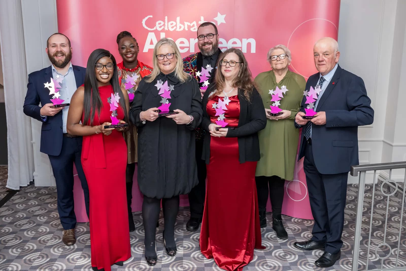 A diverse group of eight people, all dressed in formal attire, smiling and holding star-shaped awards in front of a pink banner that reads "Celebrate Aberdeen."