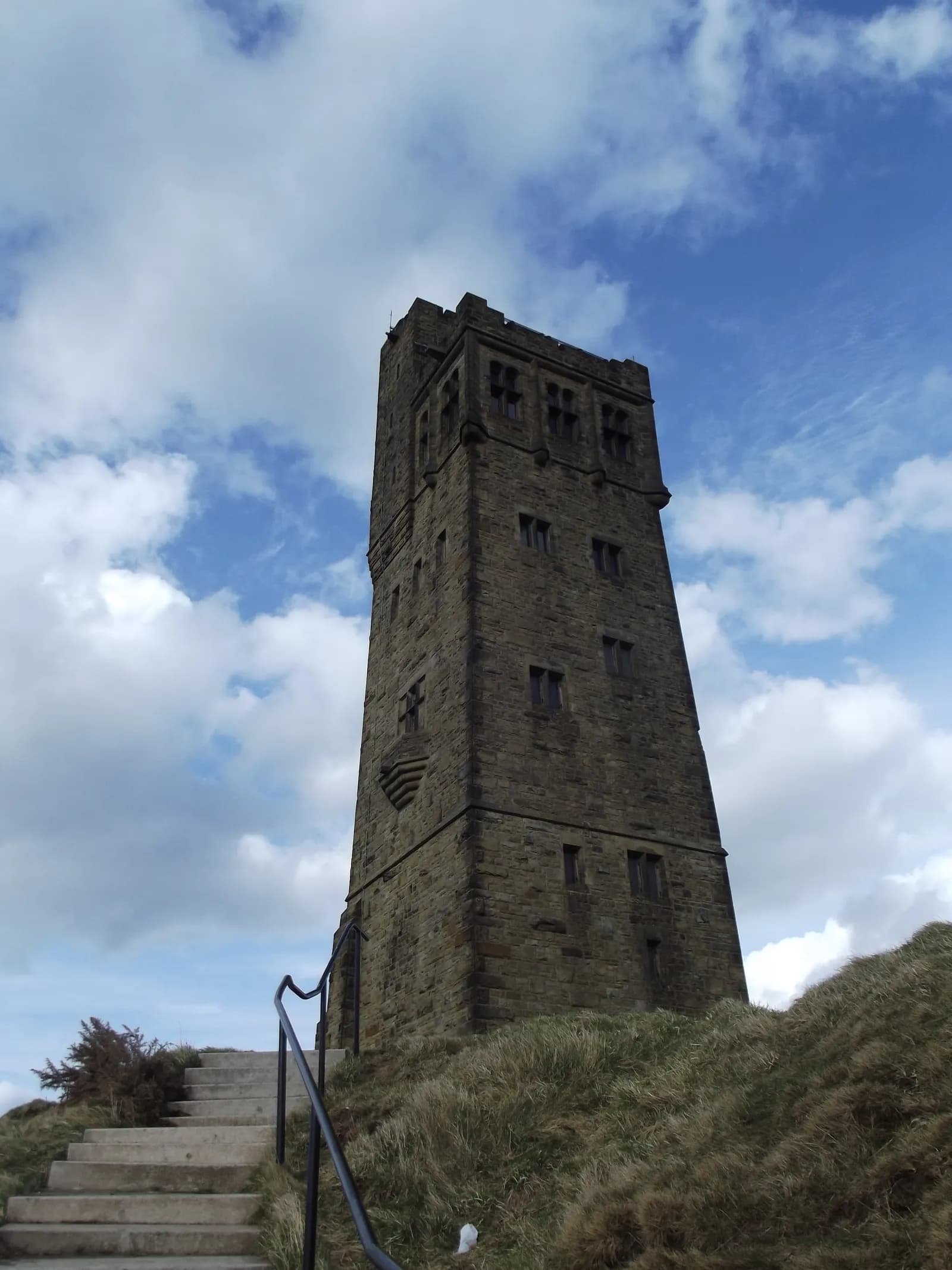 A tall, rectangular stone tower with multiple windows stands on a grassy hill under a blue sky with scattered clouds.