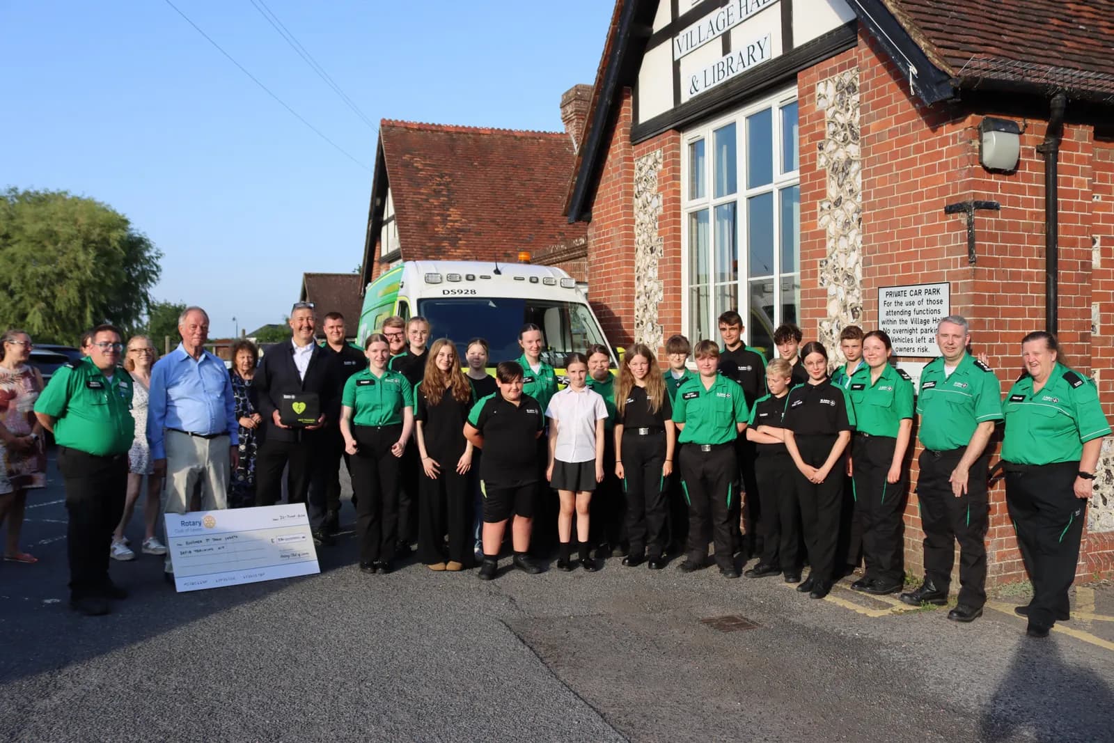 A group of adults and teenagers in green and black uniforms stand in front of a brick building and an ambulance, with a large check visible in the foreground.