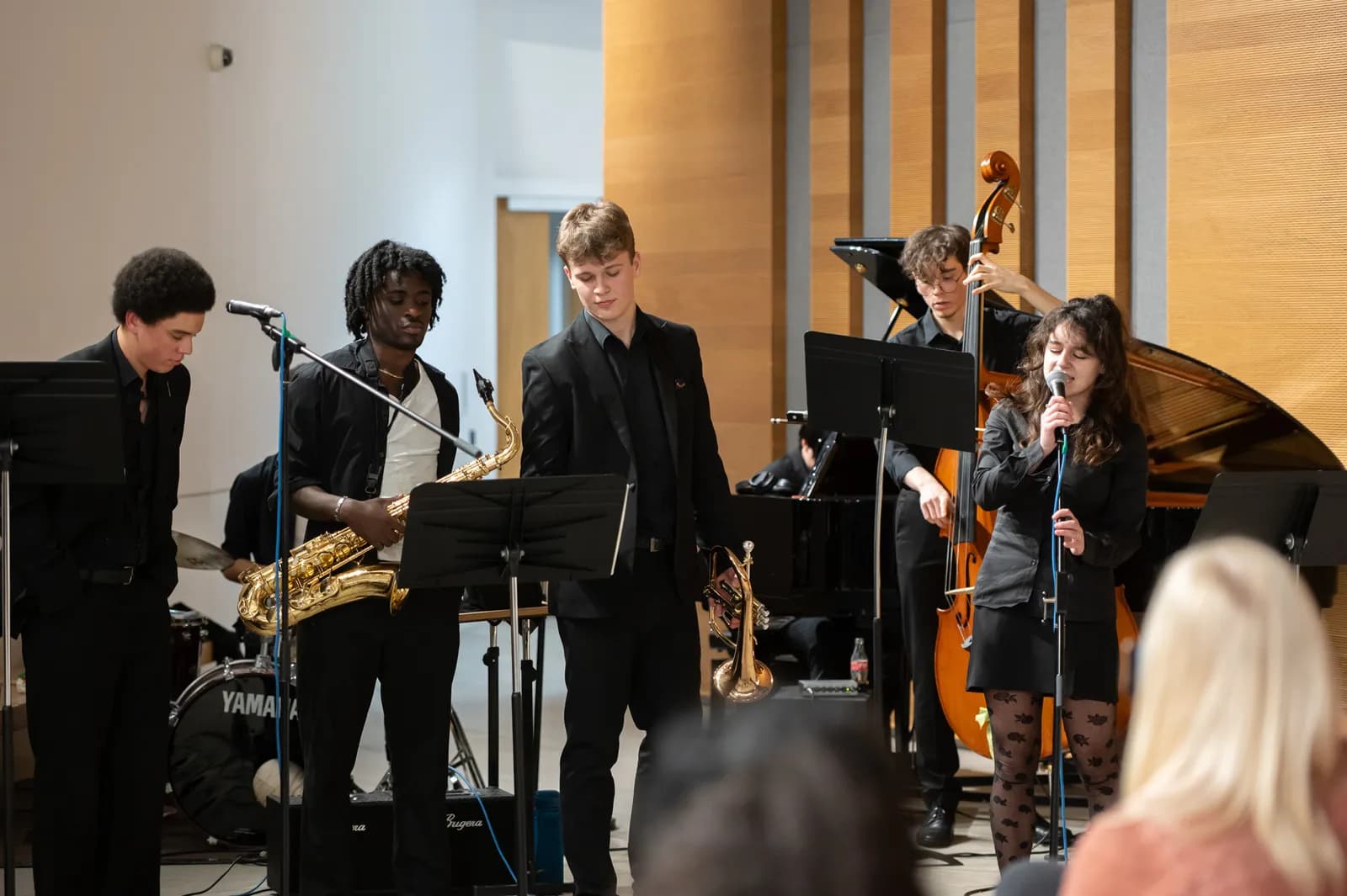 A diverse group of five young musicians, dressed in black, perform on stage with instruments including a saxophone, an upright bass, a piano, and a singer at the microphone, while an audience watches.