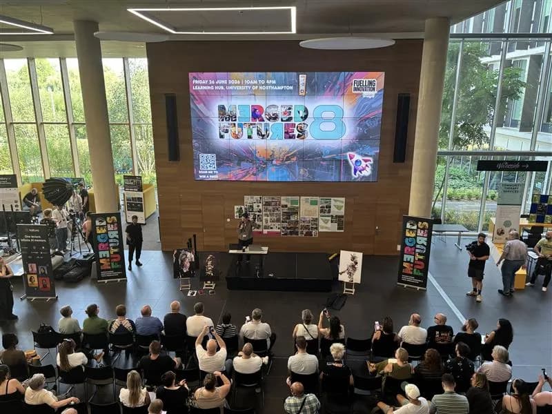 A wide shot of an indoor event with a large screen displaying "MERGED FUTURES 8," with an audience seated in front of a stage where a person is speaking, and various production equipment and staff around the room.