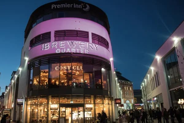 A modern building with a rounded facade illuminated with purple light and displaying 'The Brewery Quarter' in large letters, with a street full of people walking below at dusk.