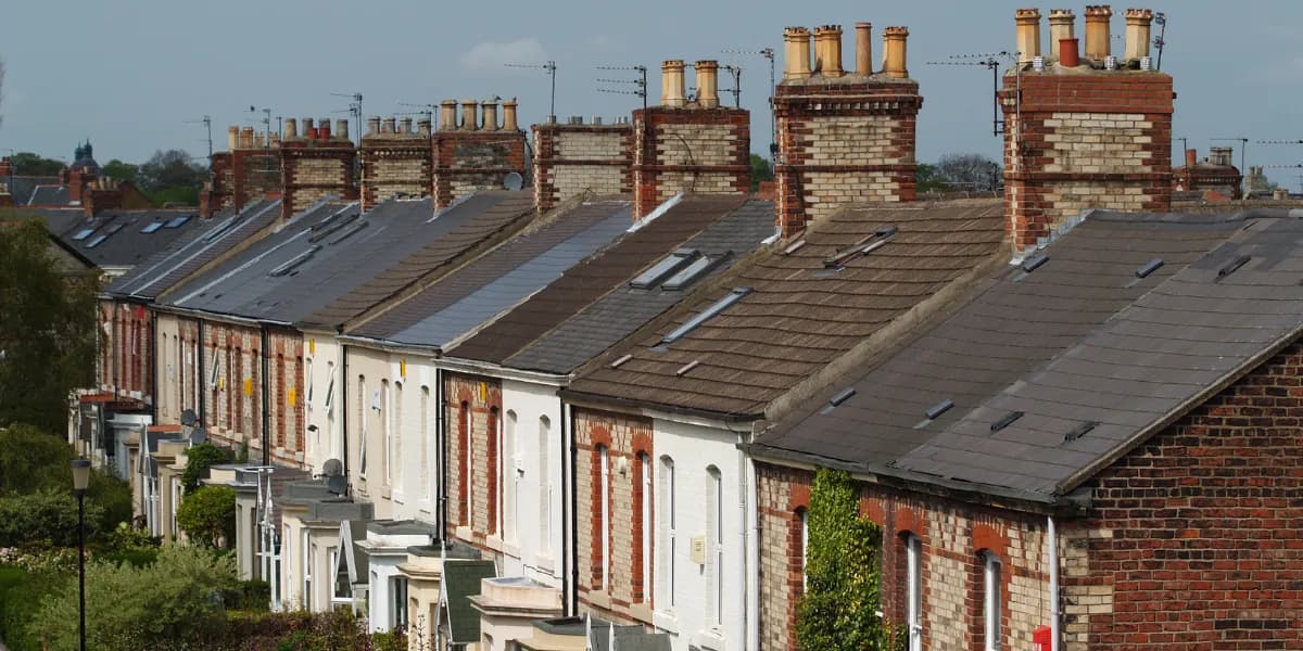 A long row of terraced houses with varying brick and painted facades, featuring an array of dark roofs with chimneys and skylights under a cloudy sky.