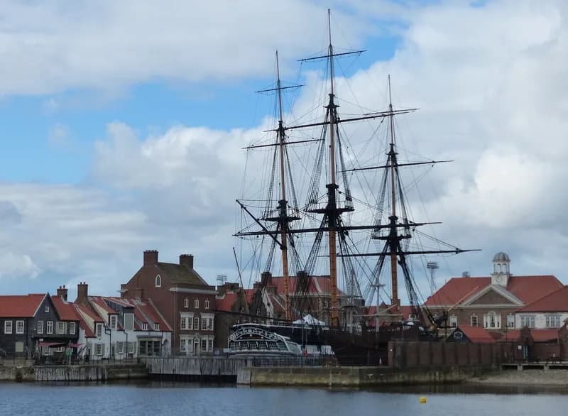 A large, multi-masted sailing ship with intricate rigging stands docked by a bustling waterfront lined with traditional Dutch-style buildings under a partly cloudy sky.