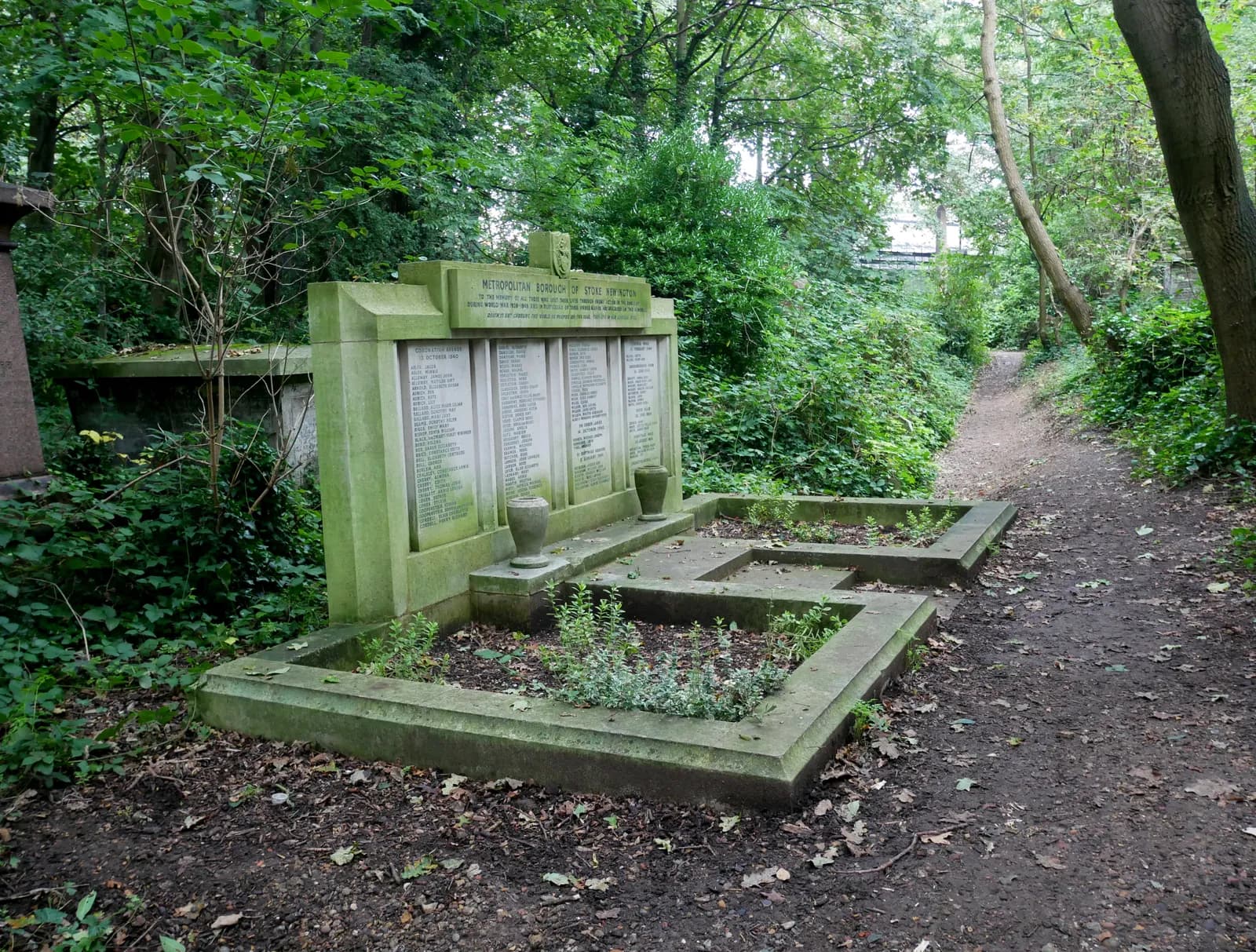 A moss-covered stone memorial with names inscribed on it stands in a wooded area with a dirt path leading into the distance.