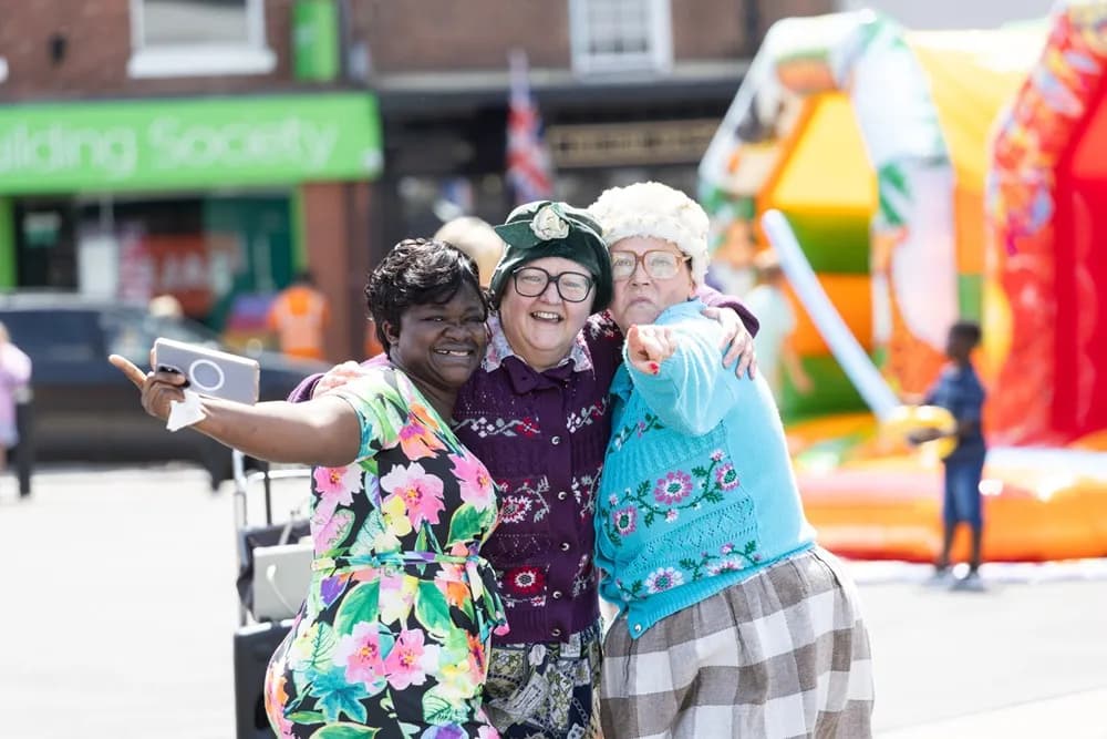 Three women in costume posing for a selfie with a blurred background of buildings and an inflatable slide.