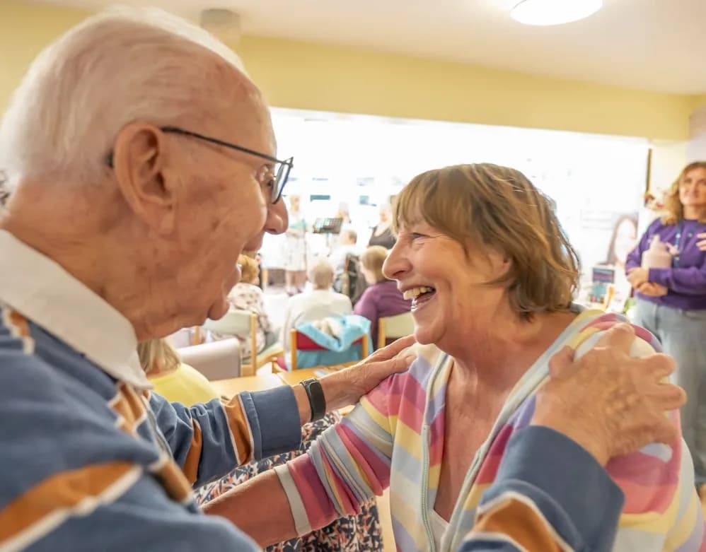 An elderly man in glasses and a collared shirt places his hands on the shoulders of an older woman in a striped top who is laughing joyfully.